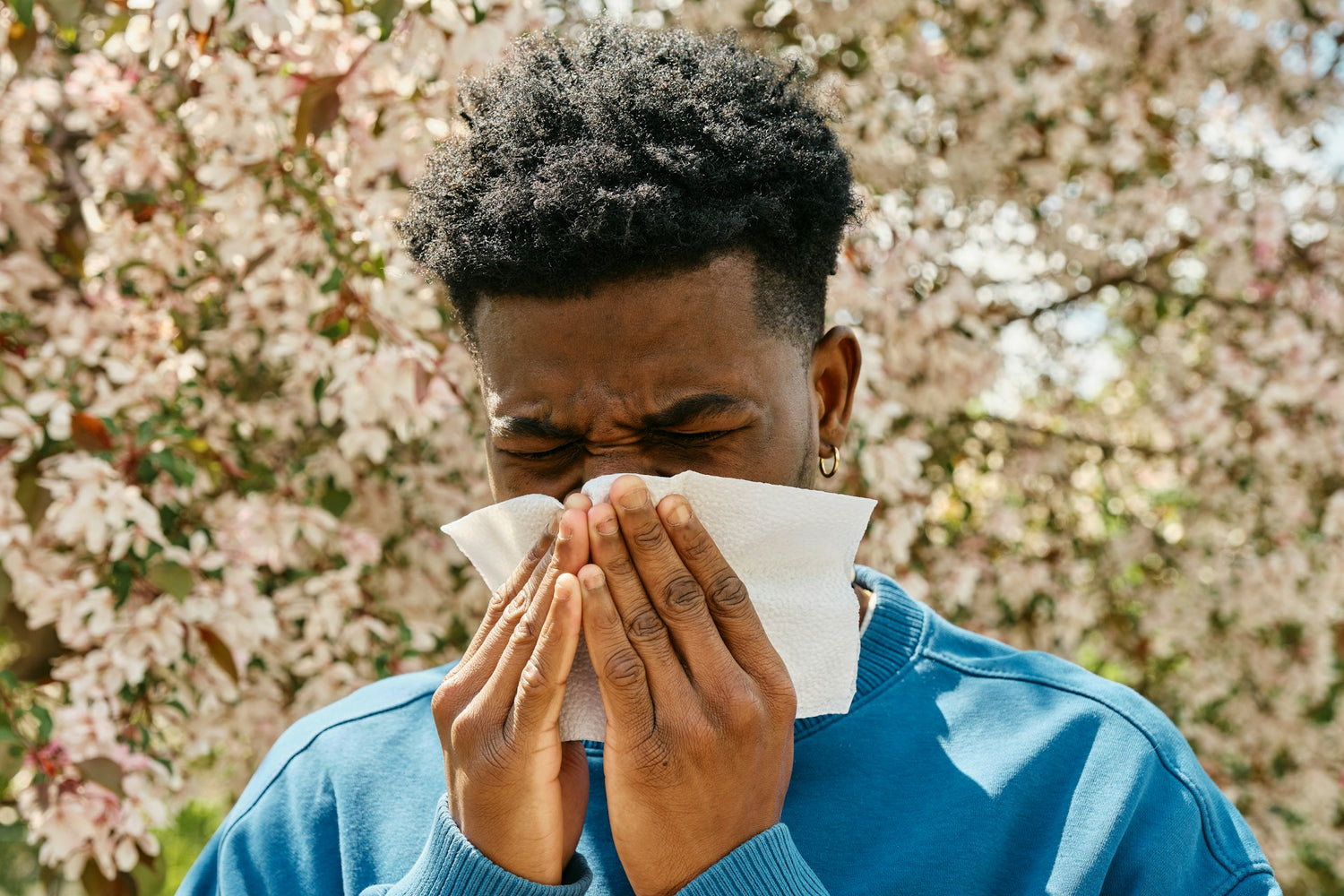 A young man outdoors sneezing into a tissue, with blooming flowers in the background, symbolising how symptoms of yeast sensitivity may overlap with allergies in certain cases.