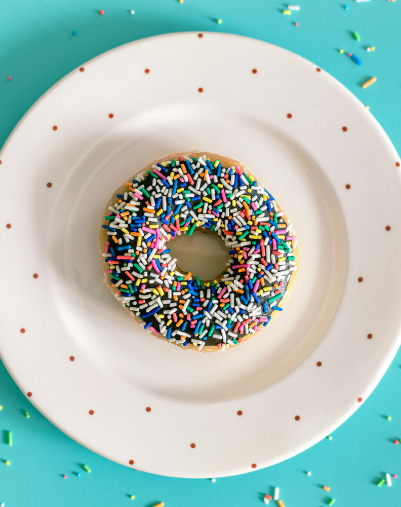 A single doughnut covered in colourful sprinkles on a white plate with polka dots, often yeast-leavened and potentially unsuitable for individuals with yeast intolerance.